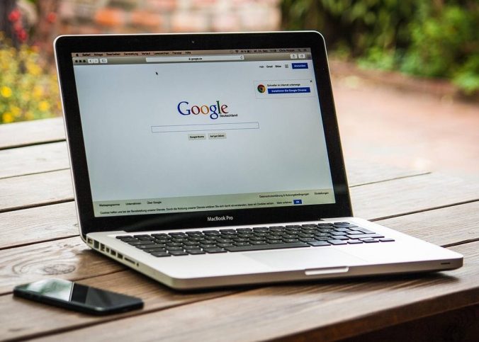 Laptop ouvert sur Google, smartphone à gauche, sur une table en bois, jardin flou