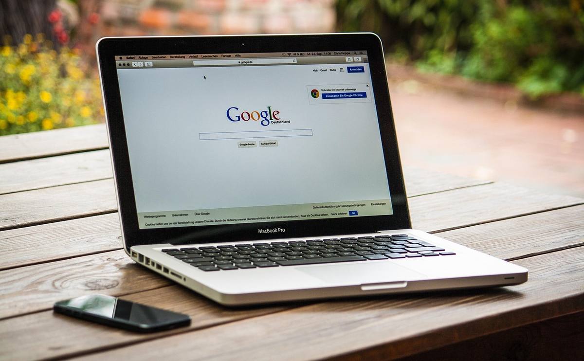 Laptop ouvert sur Google, smartphone à gauche, sur une table en bois, jardin flou